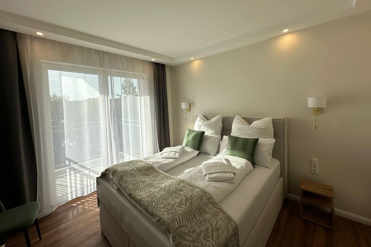 A hotel room with a bed, a small wooden side table, a wall-mounted lamp, and a view through a large window with sheer curtains.