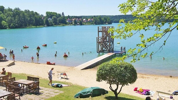 Une plage au bord d’un lac, avec une terrasse en bois, des tables de pique-nique et des parasols.