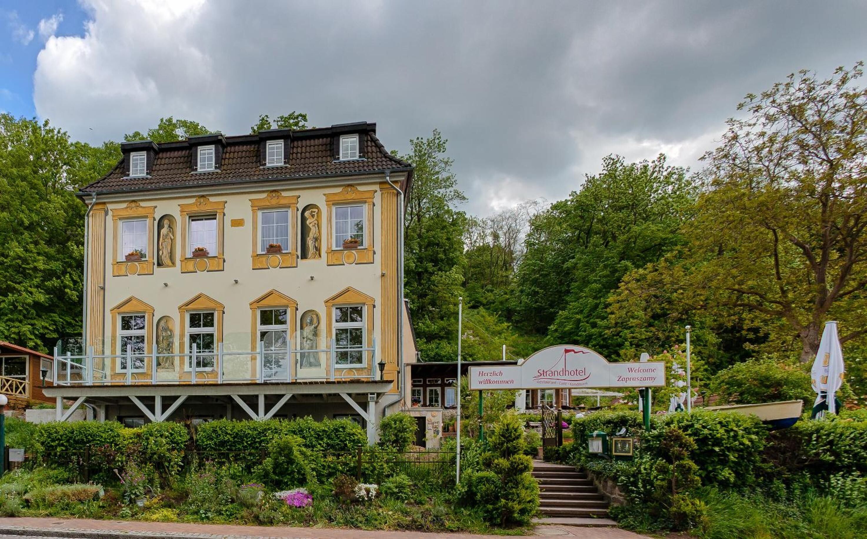 Een hotel met twee verdiepingen en een balkon, omringd door weelderige begroeiing en een bord met een gastvrije verwelkoming.