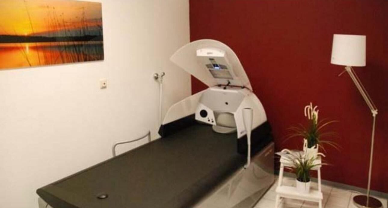 Una sala per esami medici con un lettino, un monitor a parete e un piccolo tavolo con una pianta.