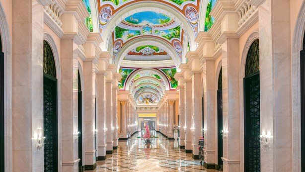 A grand hallway with marble floors and ornate ceiling decorations, featuring a central fountain and symmetrical architecture.