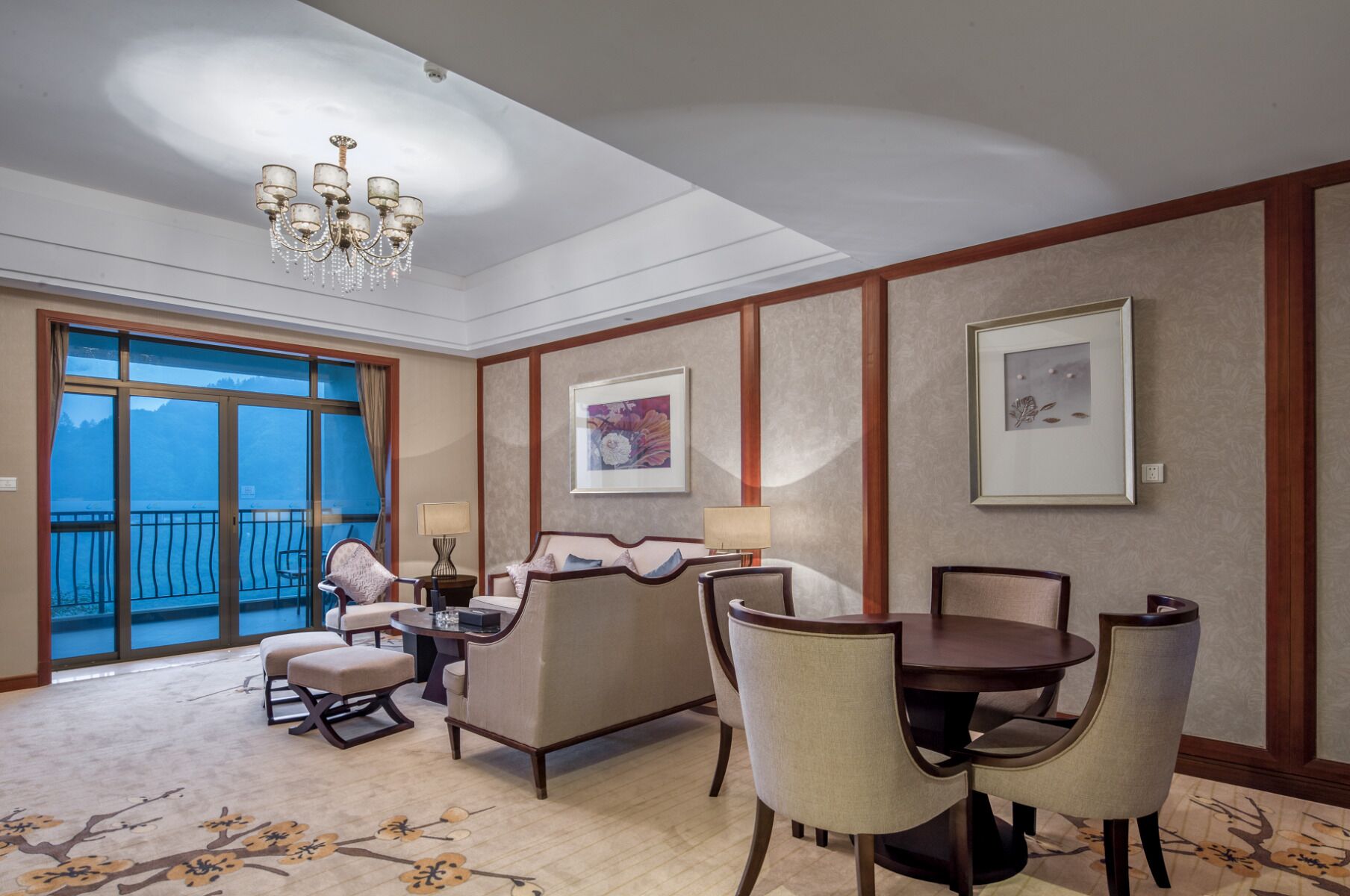 Una habitación de hotel con balcón, candelabro, un conjunto de sofás, una mesa de comedor con sillas y cuadros en las paredes.