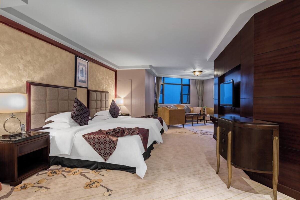 Habitación de hotel con una cama grande, un escritorio, un televisor y vistas de la ciudad al atardecer.