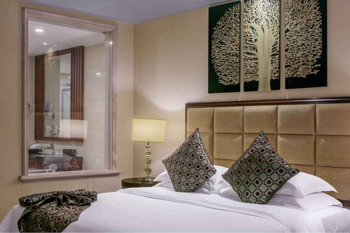 Habitación de hotel con una cama grande, un cabecero, un espejo, una mesita de noche con una lámpara y una obra de arte en la pared.