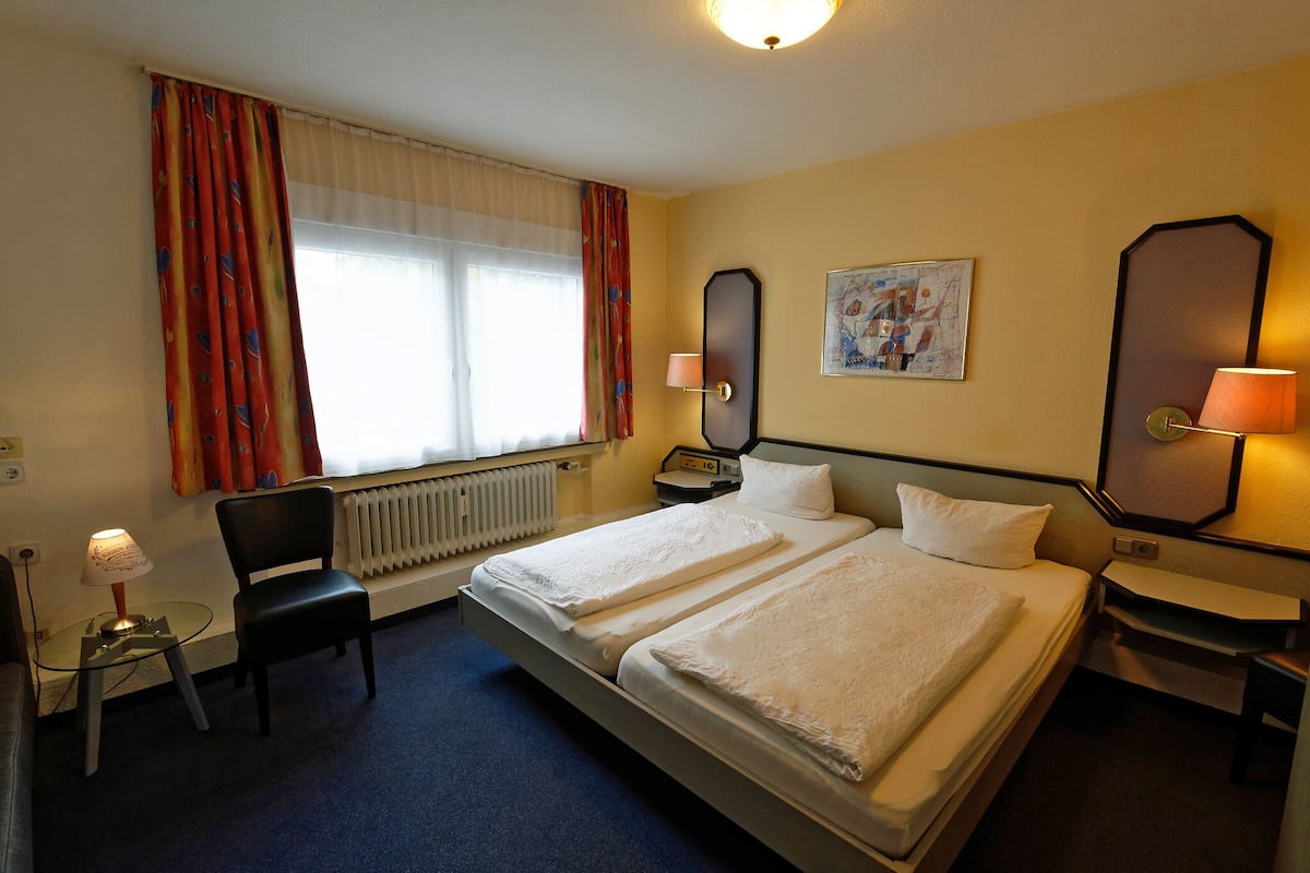 A hotel room with a double bed, two bedside tables, a chair, a desk, a lamp, a radiator, and a window with curtains.