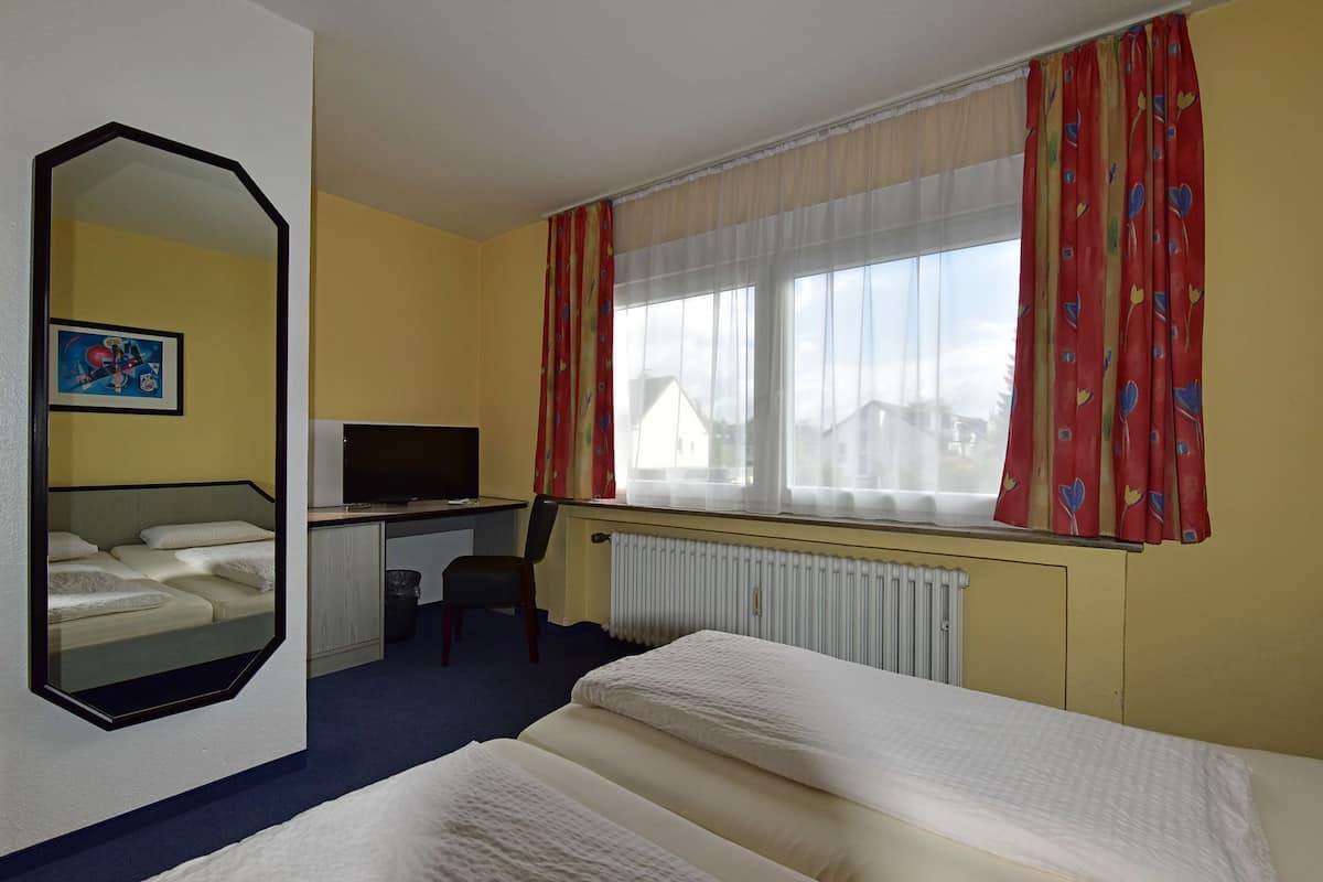 A hotel room with a bed, a desk with a television, a chair, a large mirror, and a window with curtains.