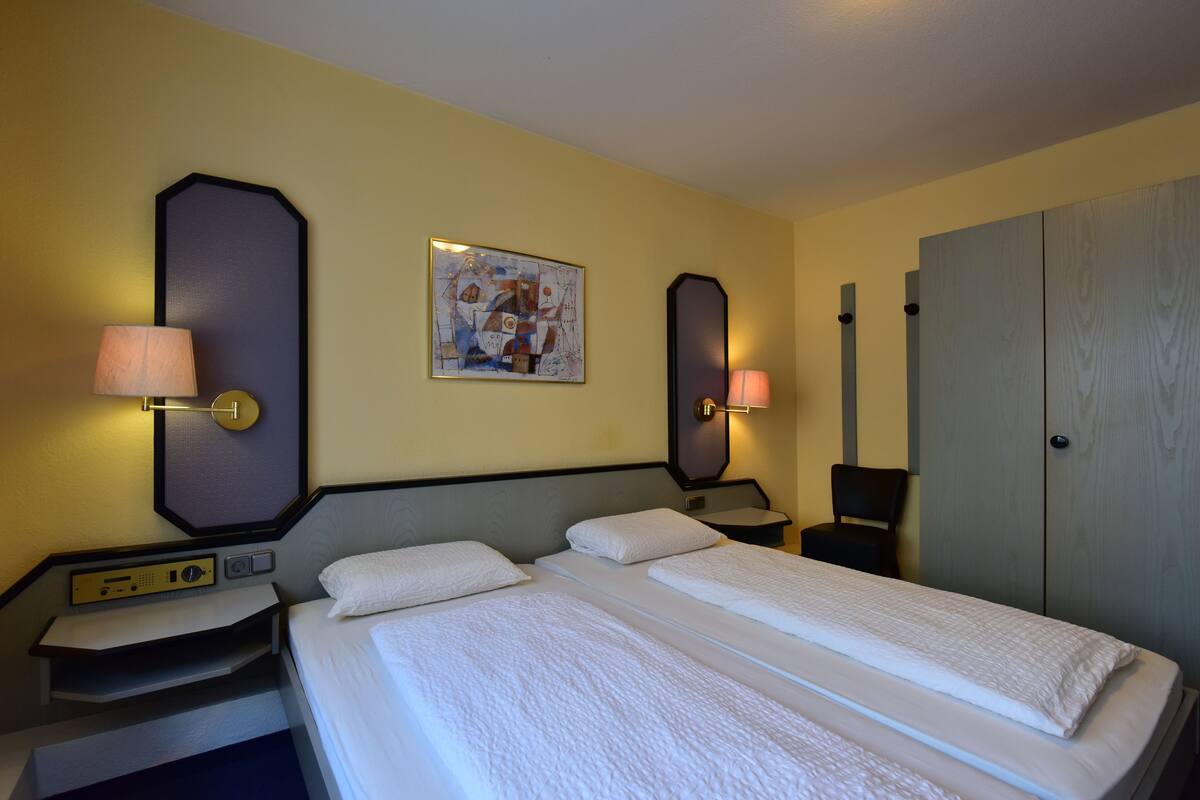 A hotel room with a double bed, two bedside tables, a desk with a lamp, a wall-mounted painting, and a wardrobe.