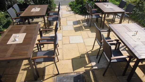 Un patio al aire libre con varias mesas y sillas de madera, dispuestas sobre un suelo de baldosas de piedra.