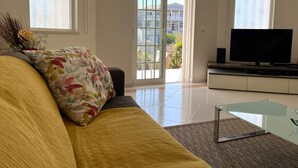 Ein Wohnzimmer mit Sofa, Glastisch, Flachbildfernseher und einem großen Fenster mit Blick ins Freie.