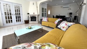 Ein Wohnzimmer mit einer Couch, einem Couchtisch und einem Kamin. Im Hintergrund befindet sich ein Essbereich mit Tisch und Stühlen.