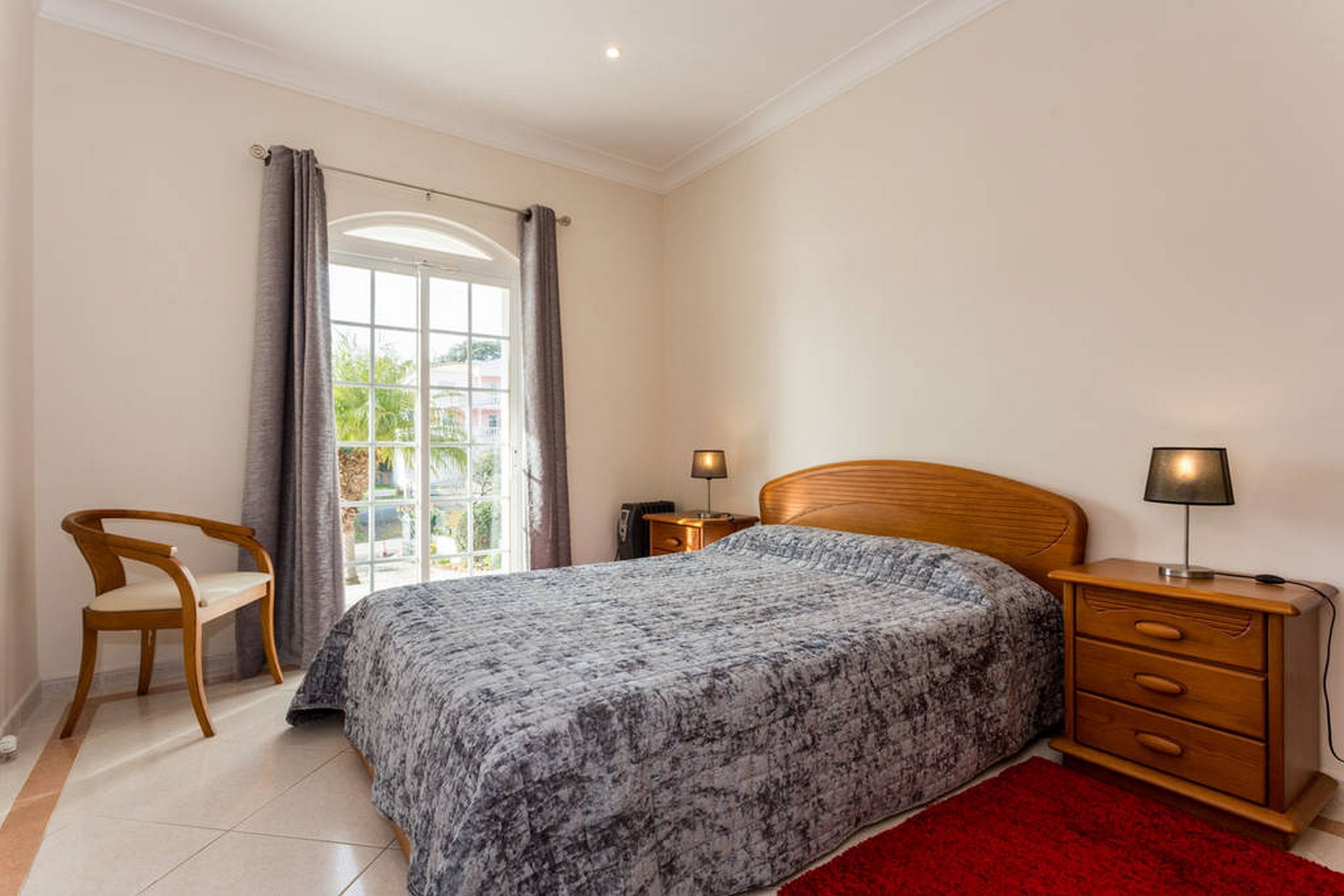 A bedroom with a bed, a wooden nightstand, a chair, a red rug, and a window with curtains.