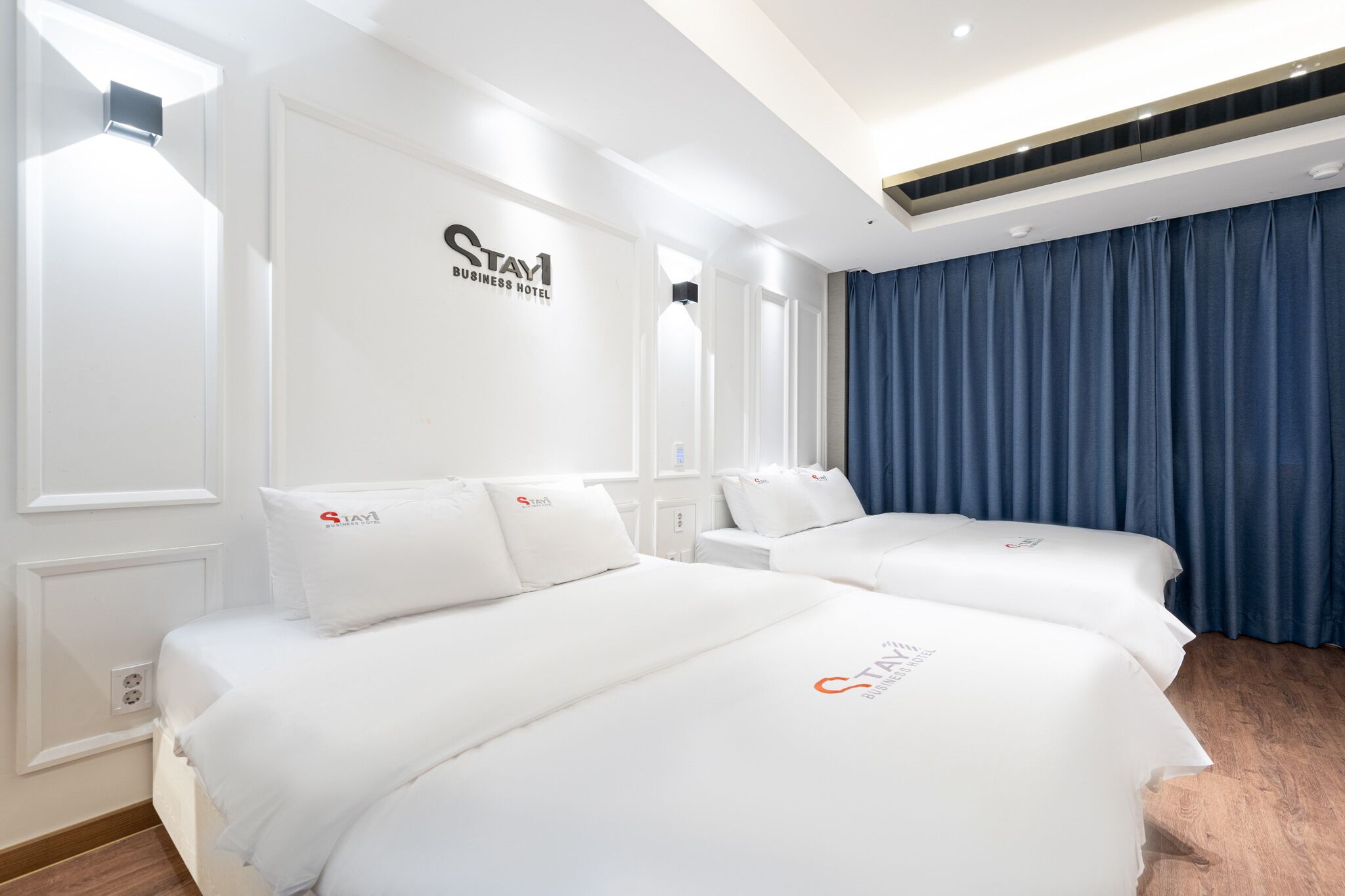 A hotel room with two beds, a large window with curtains, a wall-mounted air conditioning unit, and a ceiling with recessed lighting.