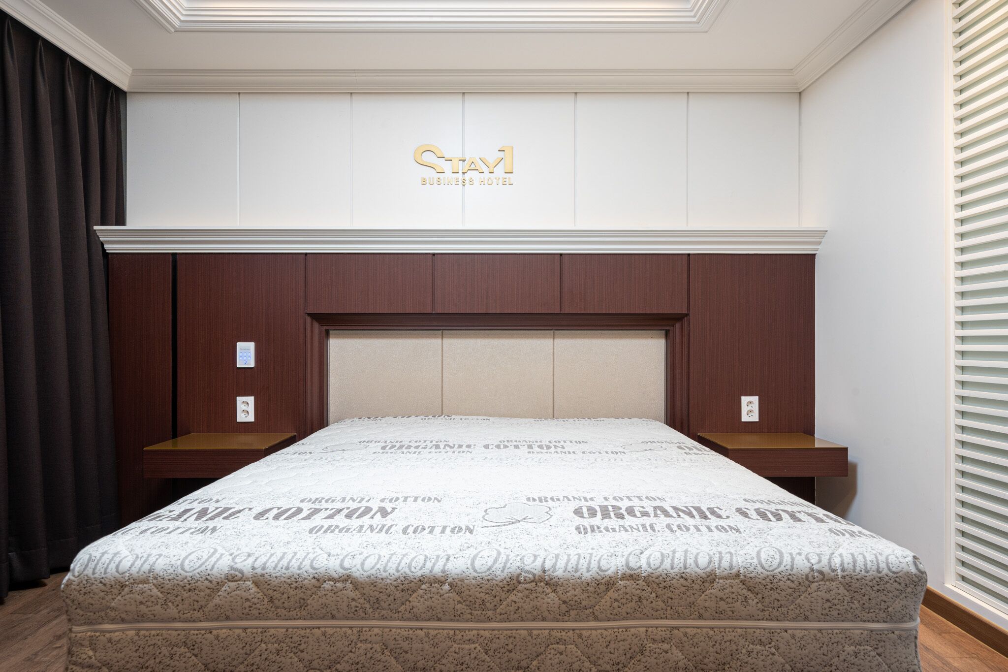 Ein Hotelzimmer mit Bett, Holz-Kopfteil und einem über dem Bett angebrachten Schild mit der Aufschrift 'Stay1 Business Hotel'.