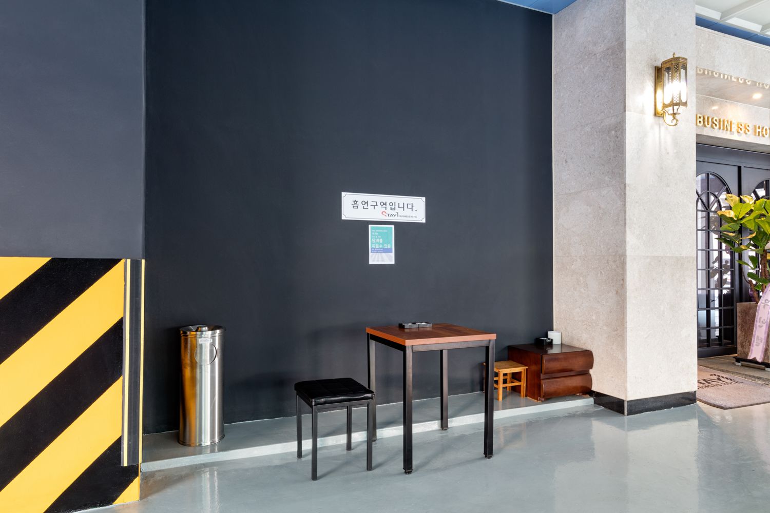 A modern hotel lobby with a dark wall, a trash bin, a small table with a chair, and a sign in Korean.