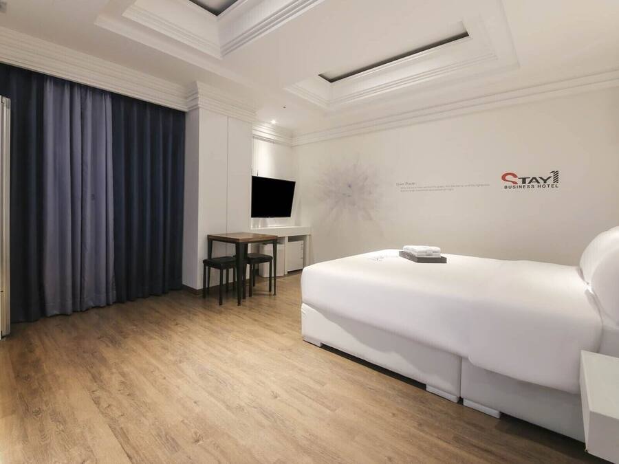 A hotel room with a white sofa, a small desk with a chair, a flat-screen TV, and a ceiling light fixture.