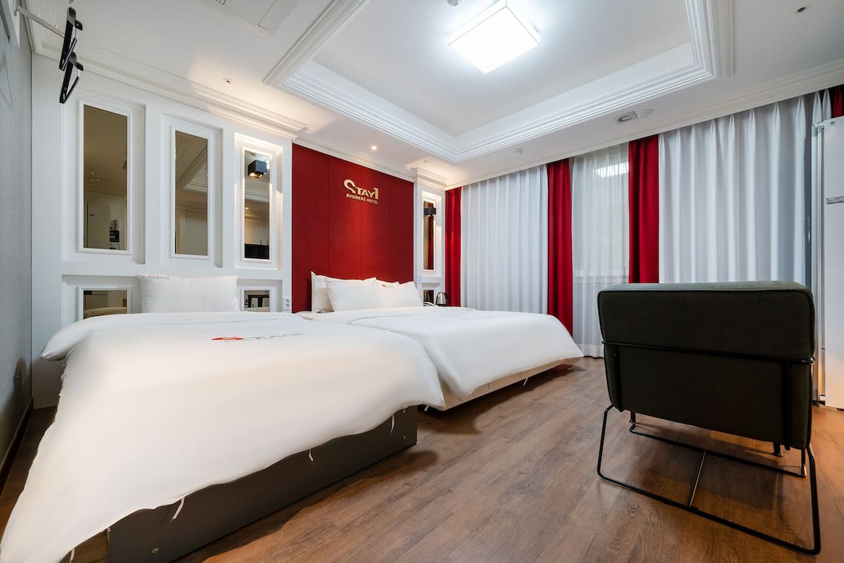 A hotel room with two beds, a red accent wall, and a black chair.