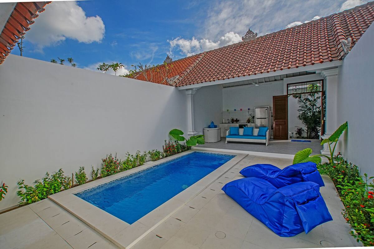 Una piscina con solárium de baldosas, un área de estar al aire libre en tonos azules y un porche cubierto con pared blanca y techo de baldosas.