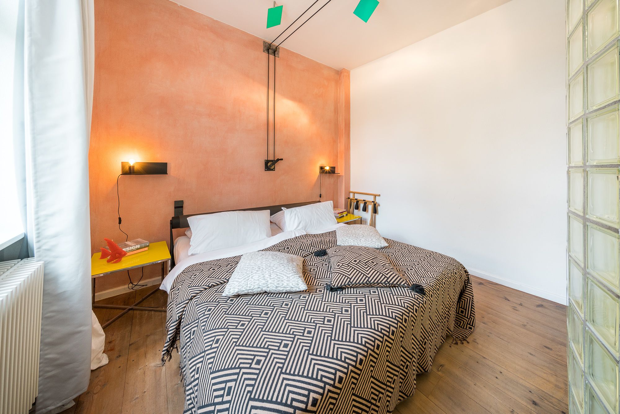 Une chambre à coucher avec un grand lit, deux tables de chevet, une commode, une lampe fixée au mur et une fenêtre avec des rideaux.