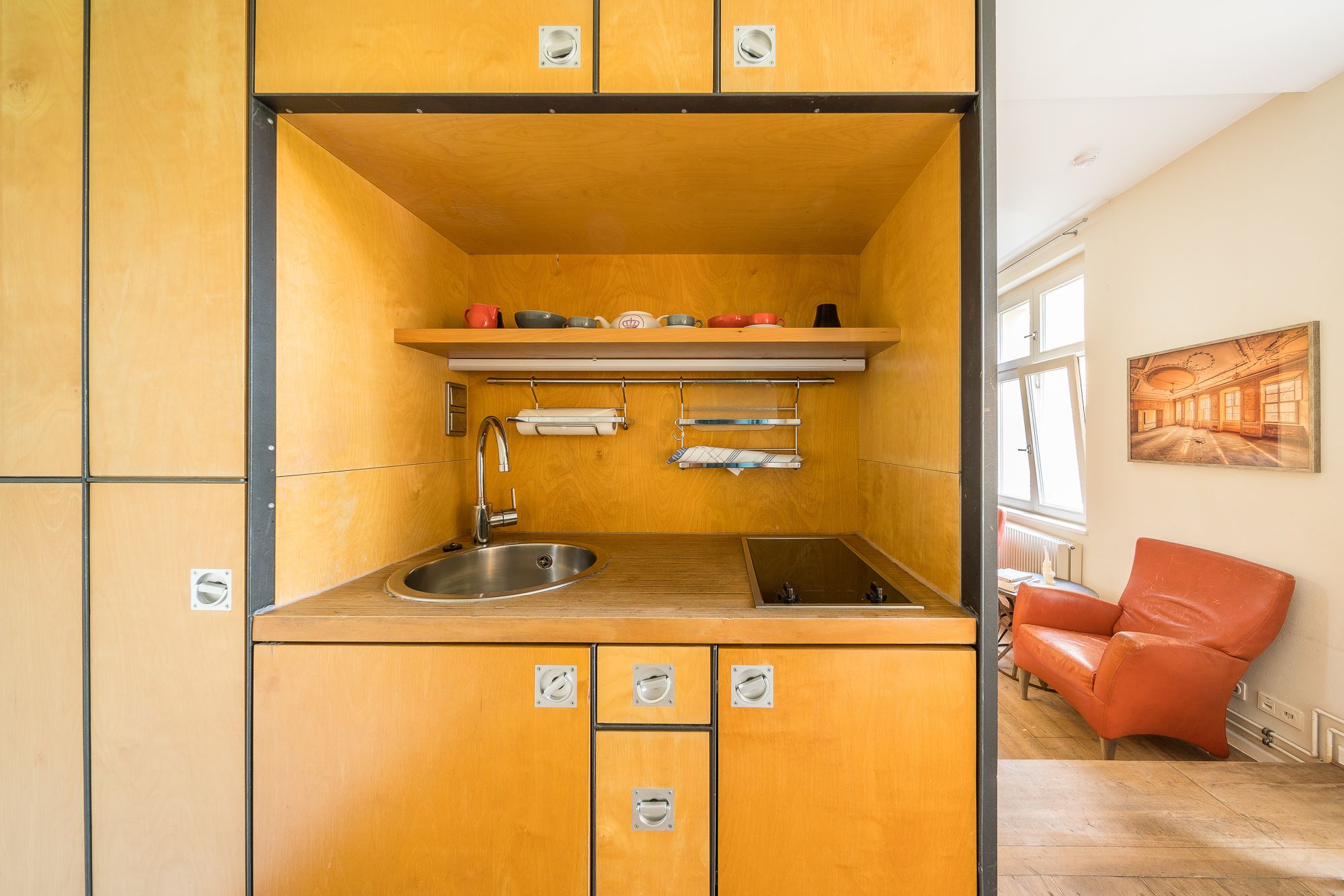 Une cuisine compacte équipée d’un évier, d’une cuisinière et d’un petit plan de travail. On y trouve une chaise en cuir rouge et un tableau accroché au mur.
