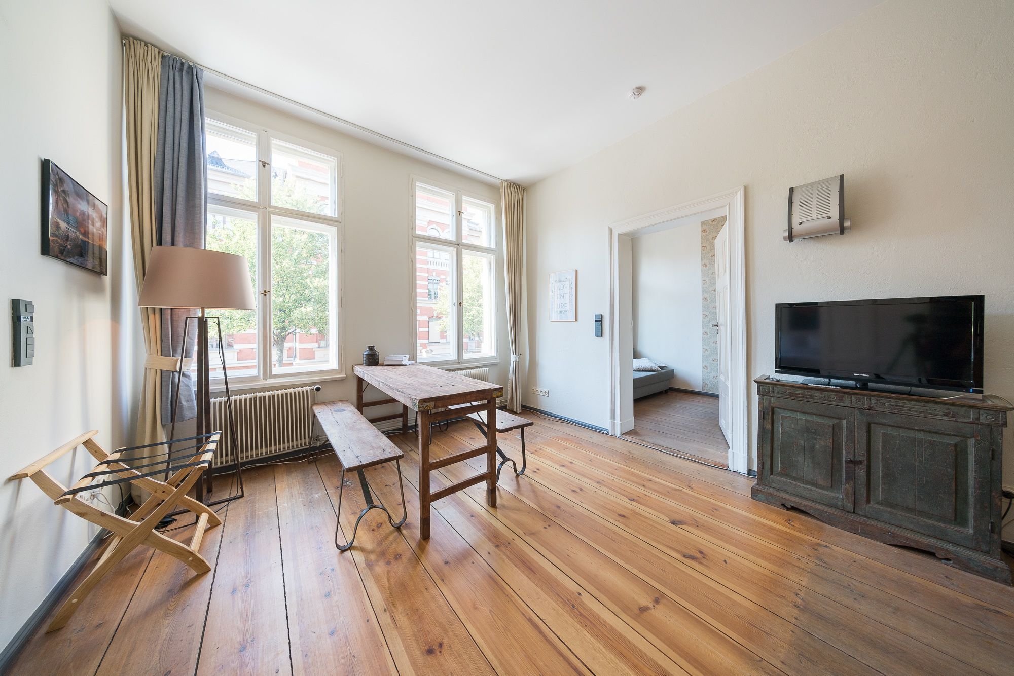 Una habitación amplia con piso de madera, un ventanal grande, una mesa, un banco, un radiador, un televisor sobre un mueble y una puerta que da a otra habitación.