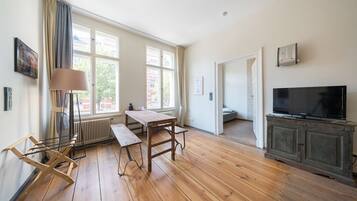 Una habitación amplia con piso de madera, un ventanal grande, una mesa, un banco, un radiador, un televisor sobre un mueble y una puerta que da a otra habitación.
