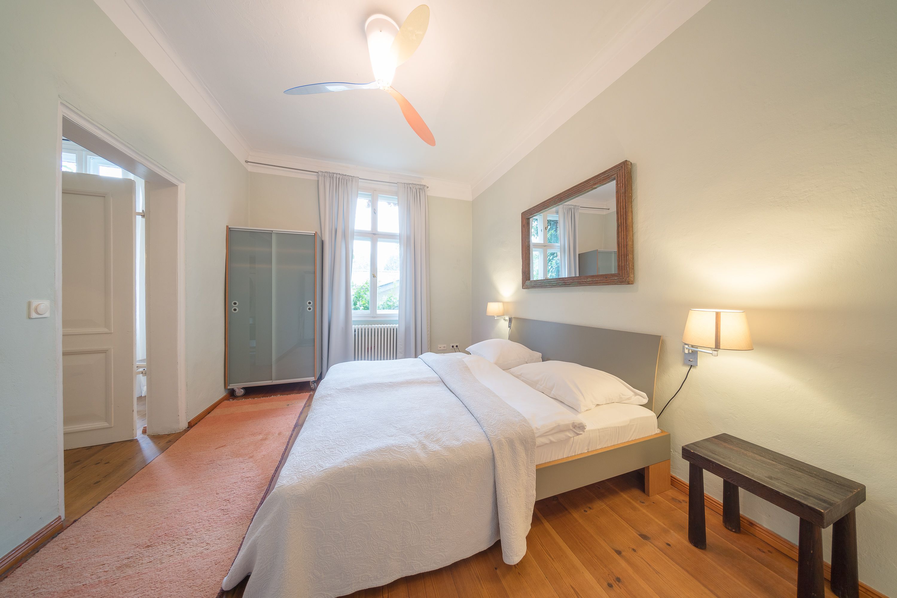 A bedroom with a bed, a bedside table, a ceiling fan, a window with curtains, and a mirror on the wall.