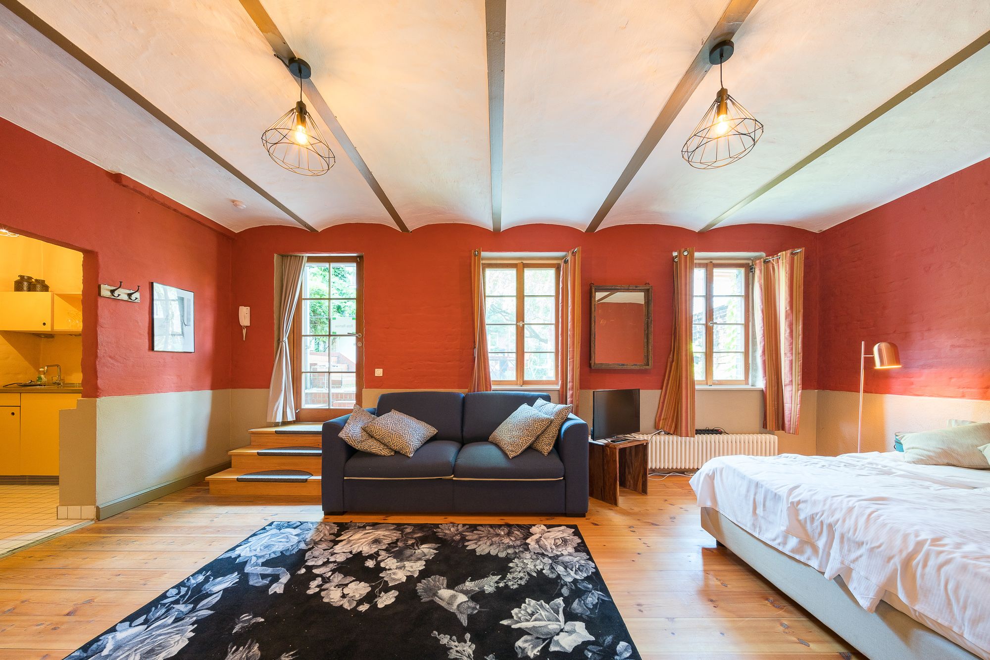 Una habitación con sofá, cama, piso de madera y ventana con cortinas.