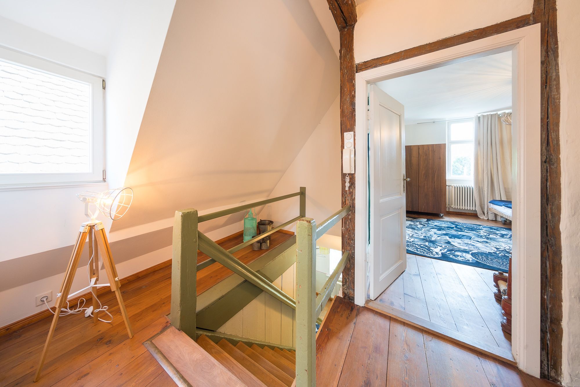 A room with a wooden staircase, a window with blinds, and a view into another room with a patterned rug.