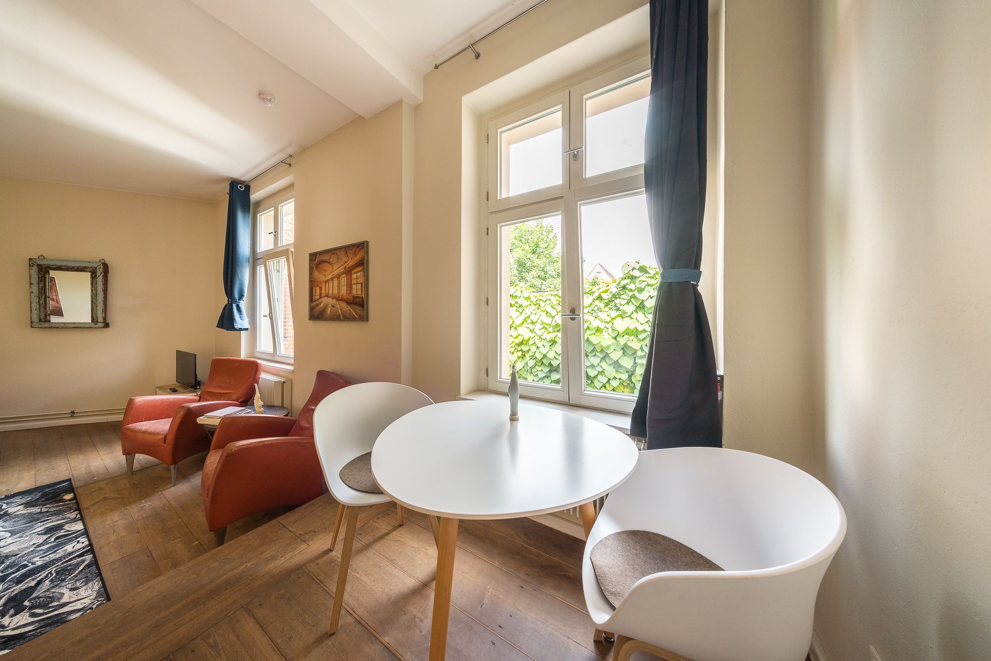 Una habitación con una mesa de comedor, dos sillas blancas, un sofá rojo y un ventanal con cortinas.
