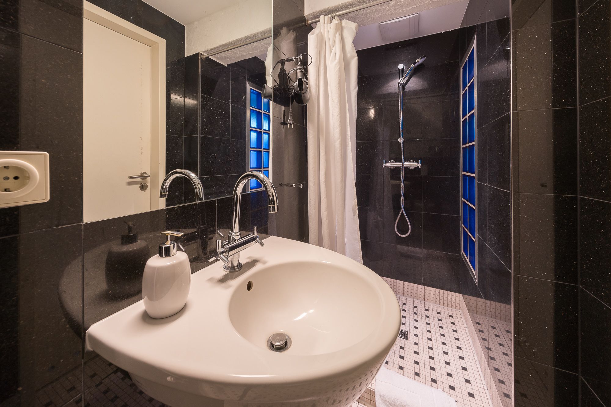 Un baño con un lavamanos blanco, un espejo, una ducha con puerta de vidrio azul y paredes de azulejos negros.
