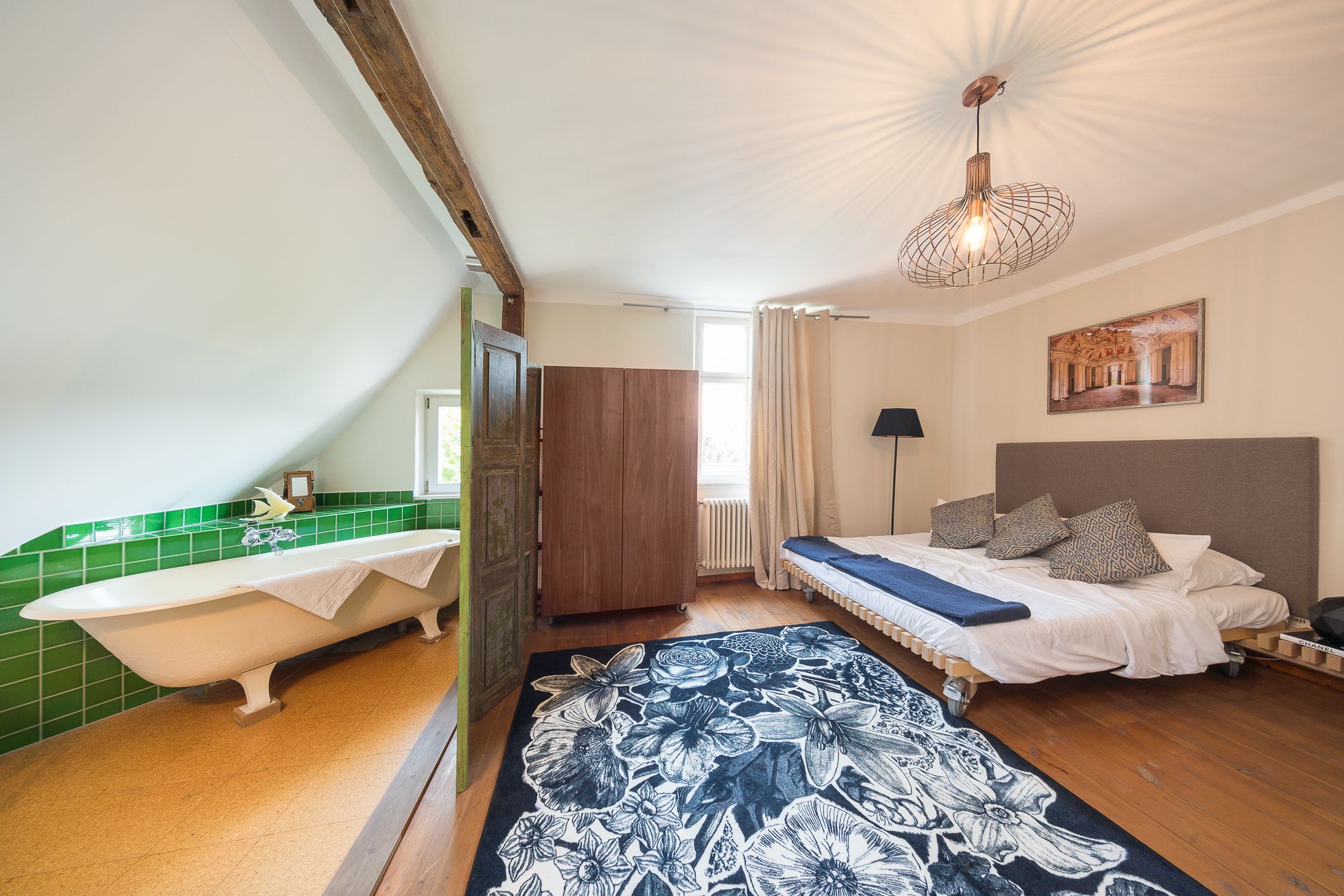 A bedroom with a bed, a nightstand, a lamp, a window, and a bathroom with a bathtub and green tiles.