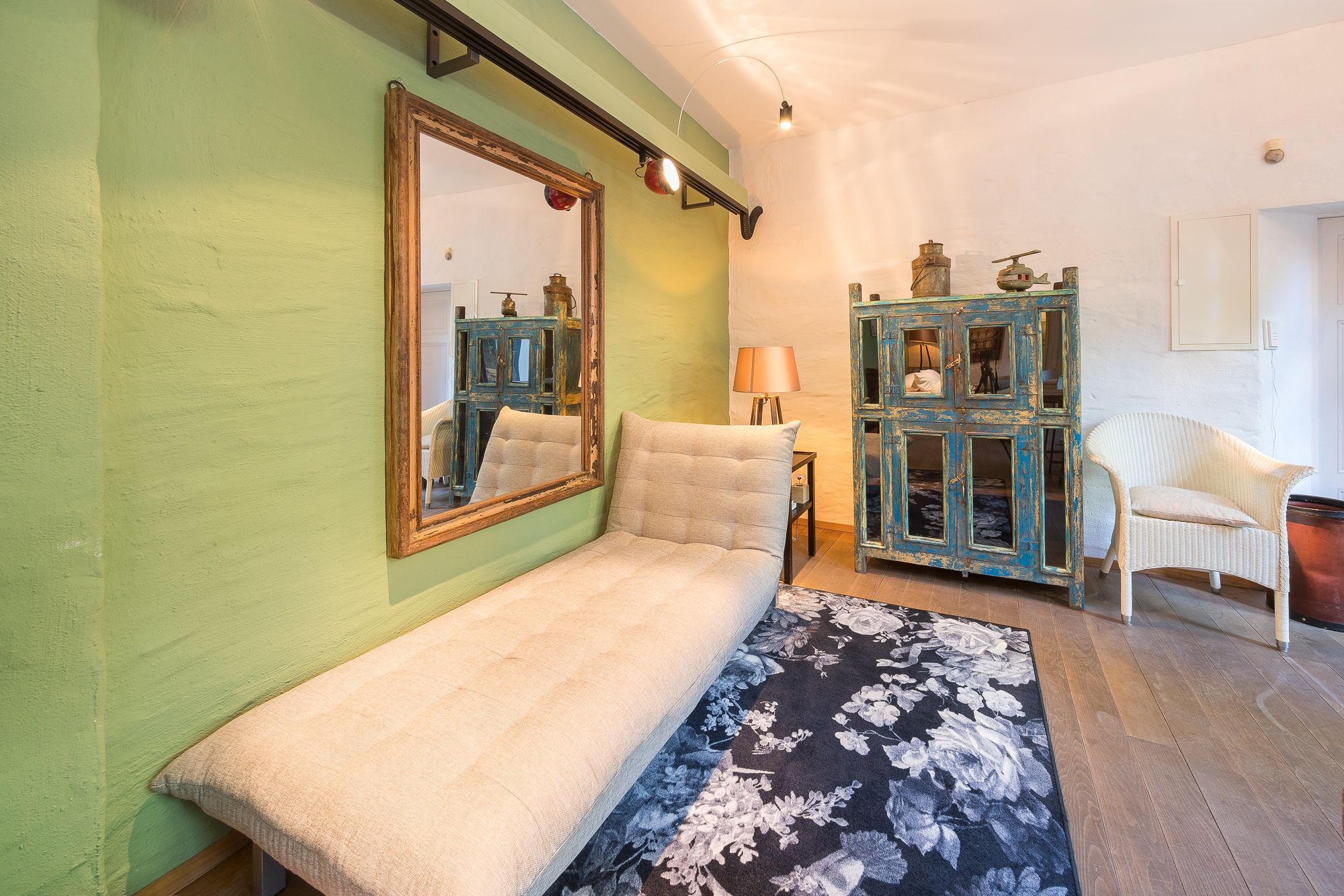 Ein Zimmer mit grüner Wand, einem großen Spiegel, einer Couch, einem Schrank mit Spiegeln, einer Lampe und einem Korbstuhl.