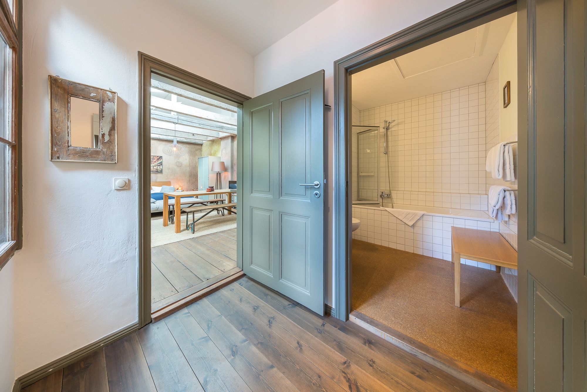 A hallway leading to a bedroom and bathroom with a wooden floor, a bed, a desk, and a shower area.
