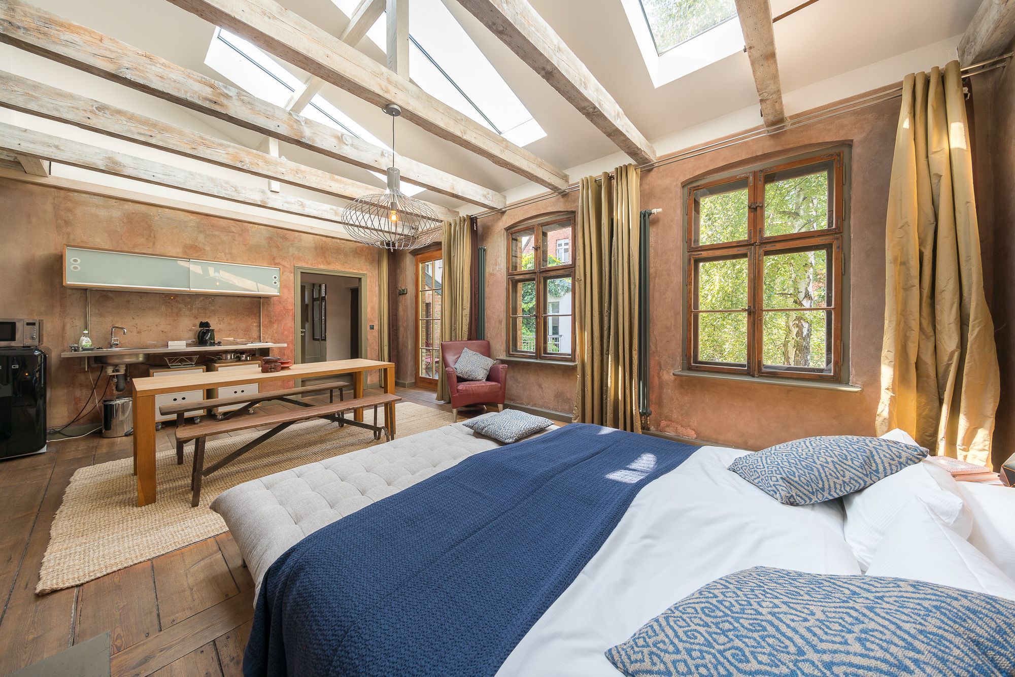 A rustic bedroom with a large bed, a wooden bench, a desk with a mirror, and a window with curtains.