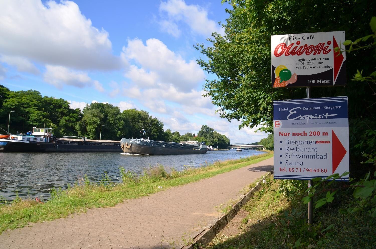 A riverside walkway with signs for Eis-Café and Exquisit hotel, including amenities like a restaurant, bistro, swimming pool, and sauna.