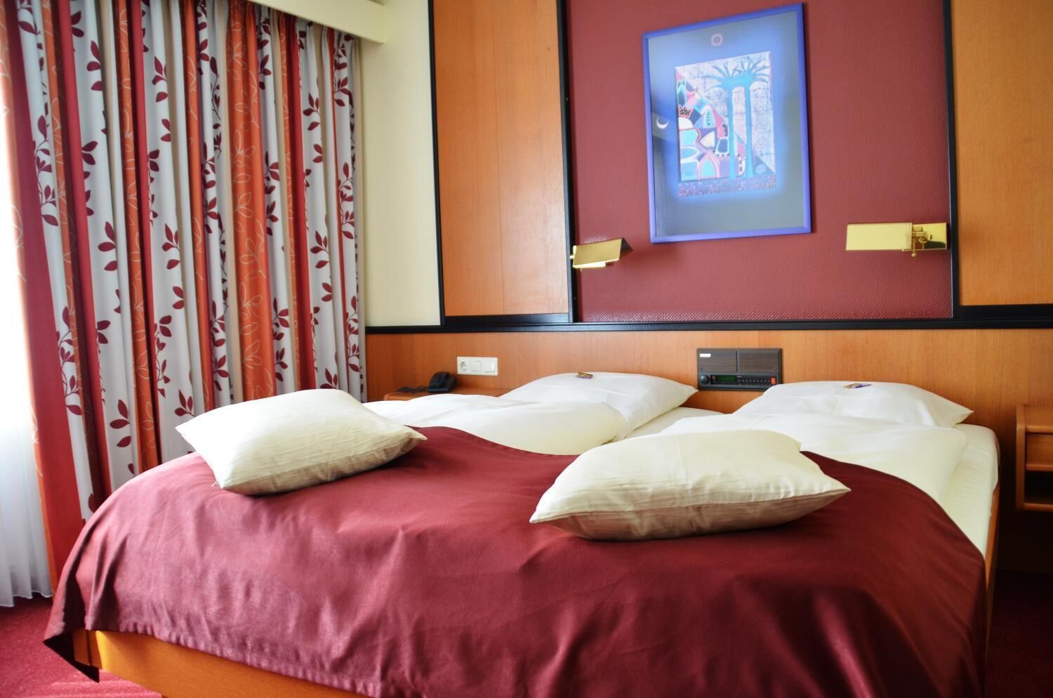 Hotellihuone, jossa on suuri sänky, kaksi yöpöytää, seinälle asennettu radio ja sängyn yläpuolella oleva kehystetty taulu.
