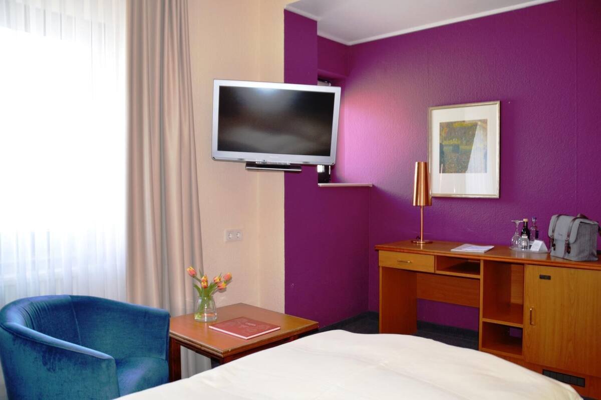 A hotel room with a bed, a desk, a chair, a television, and a painting on the wall.