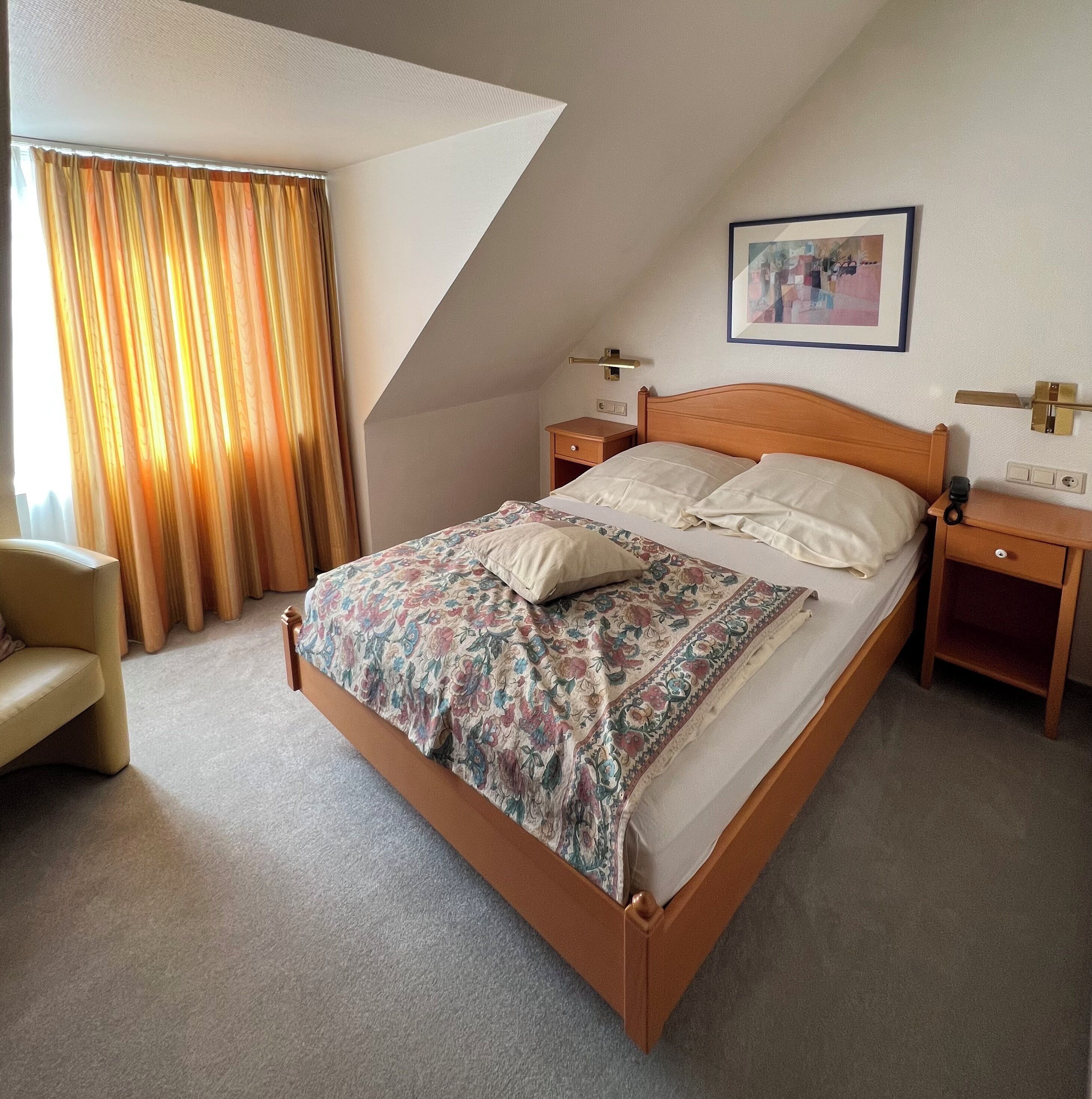 A hotel room with a single bed, a nightstand, a chair, and a painting on the wall.