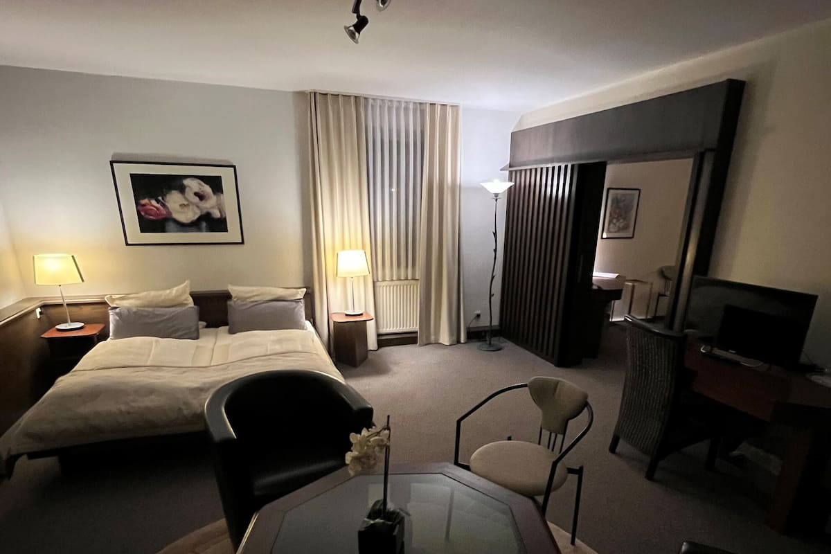 A hotel room with a bed, two bedside tables with lamps, a desk with a chair, a TV, and a large window with curtains.