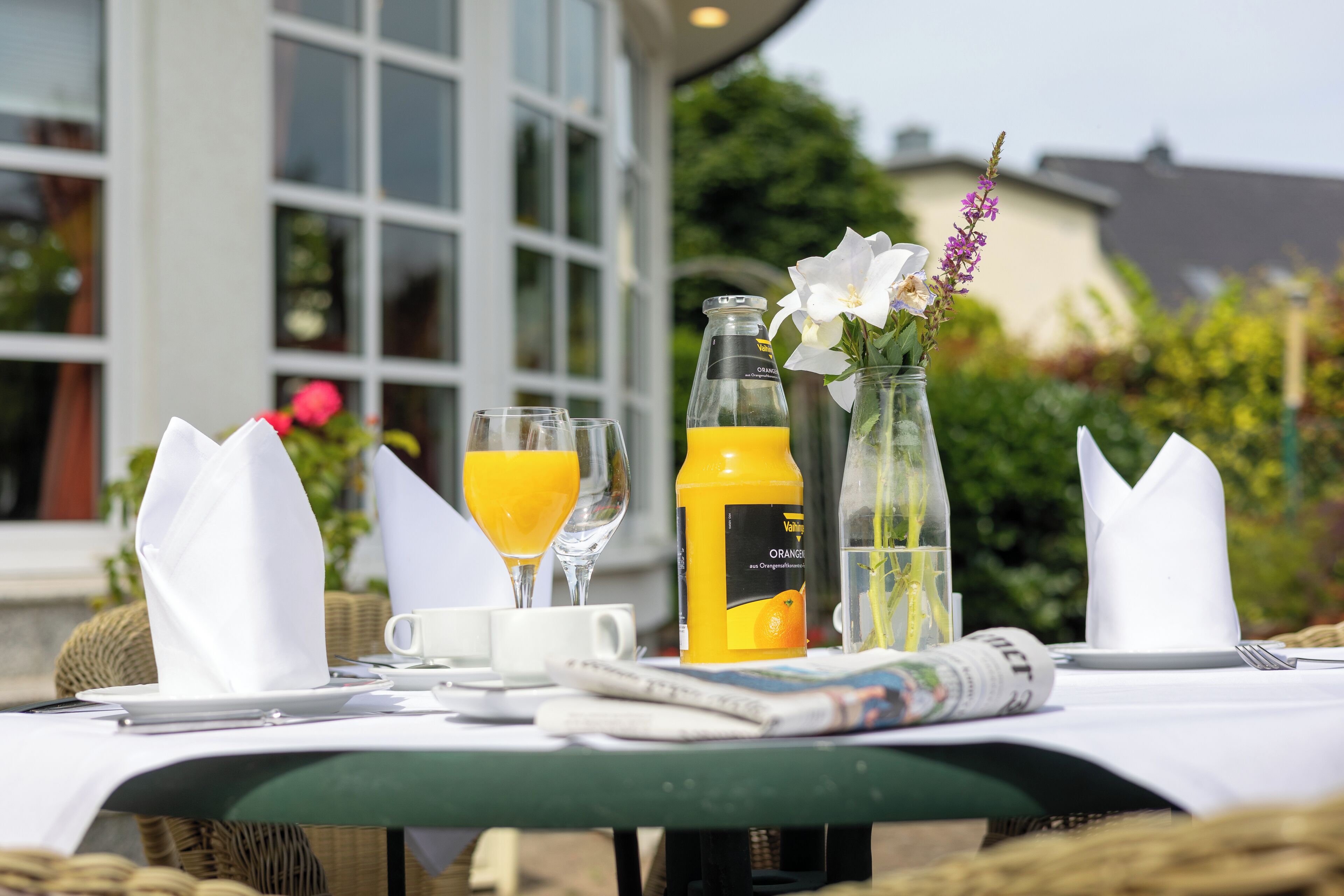 Een ontbijttafel met een fles en glas sinaasappelsap, een krant en een vaas met bloemen.