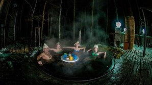 A group of people in a hot tub surrounded by a rustic wooden deck and a brick structure, with steam rising from the water.
