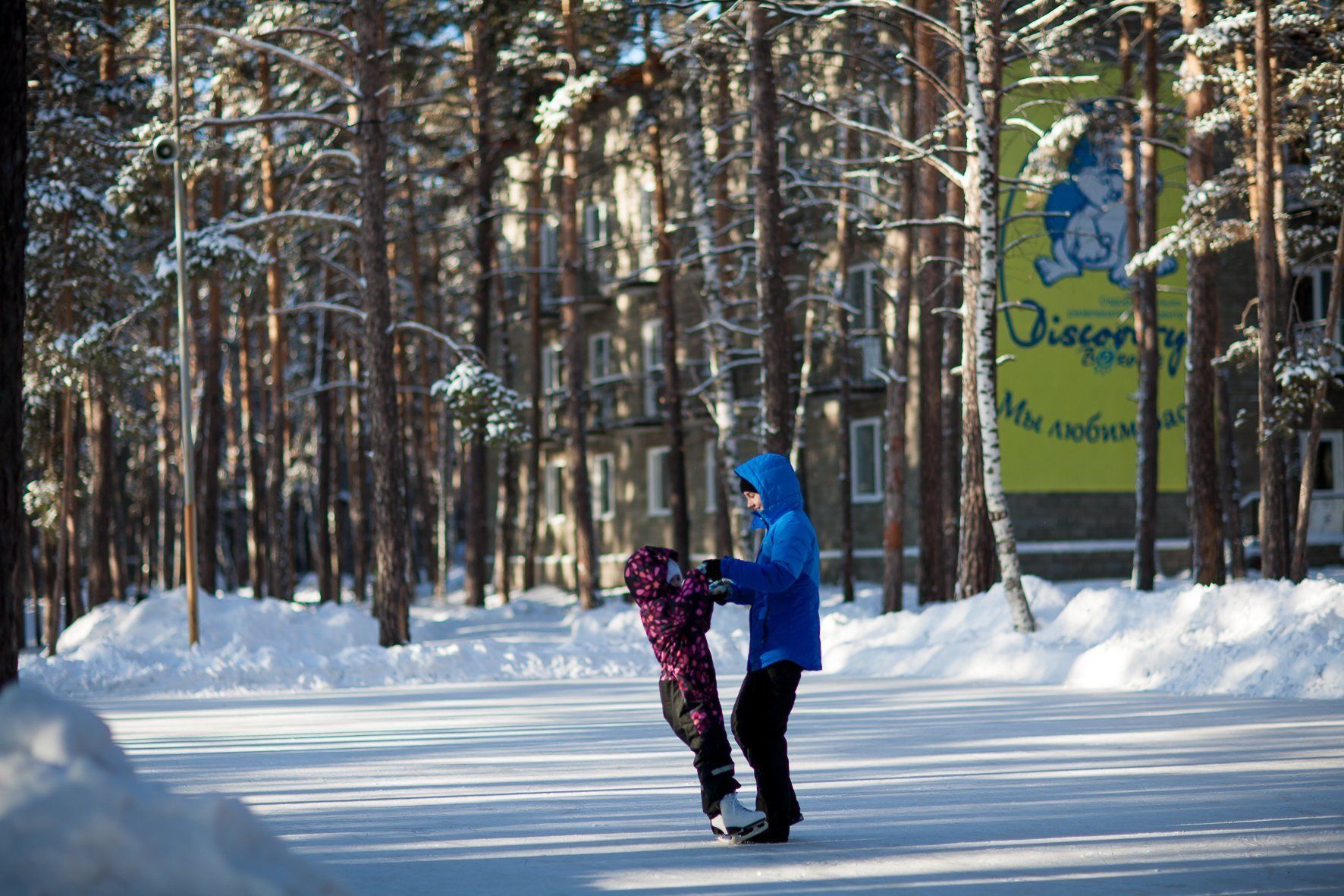 A person ice skating on a snow-covered path with trees and a building in the background.