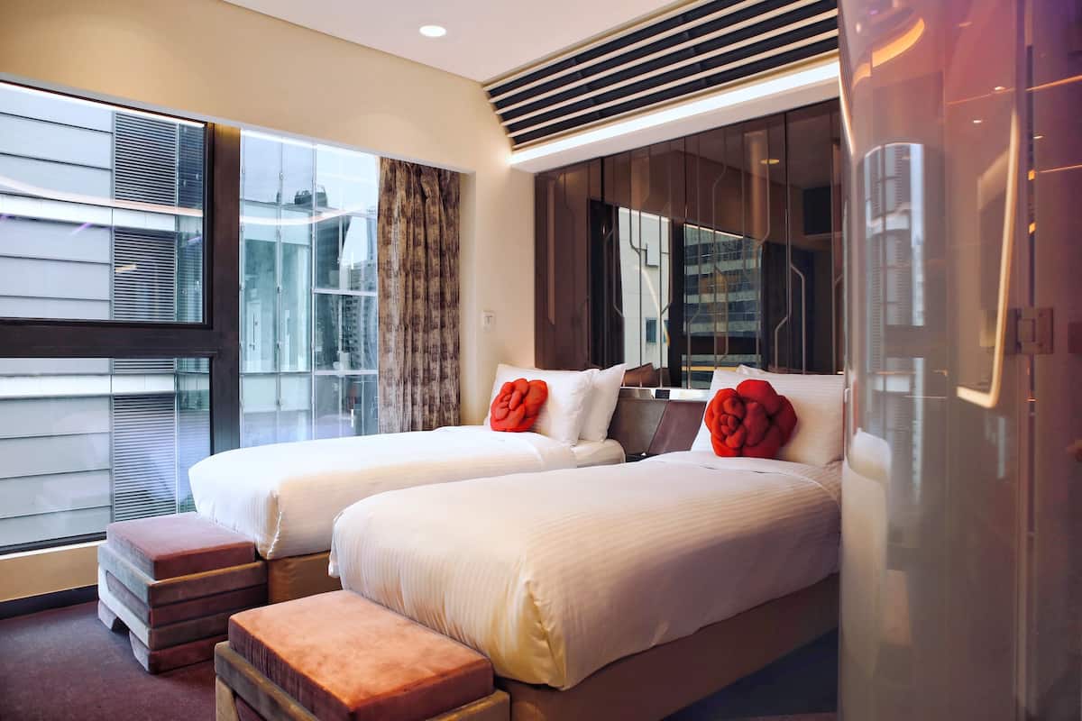 A modern hotel room with two beds, a large window with blinds, a built-in heater, and a view of the city.