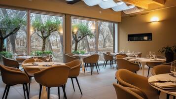 A restaurant with round tables and chairs, large windows with a view of trees, and a wooden ceiling.