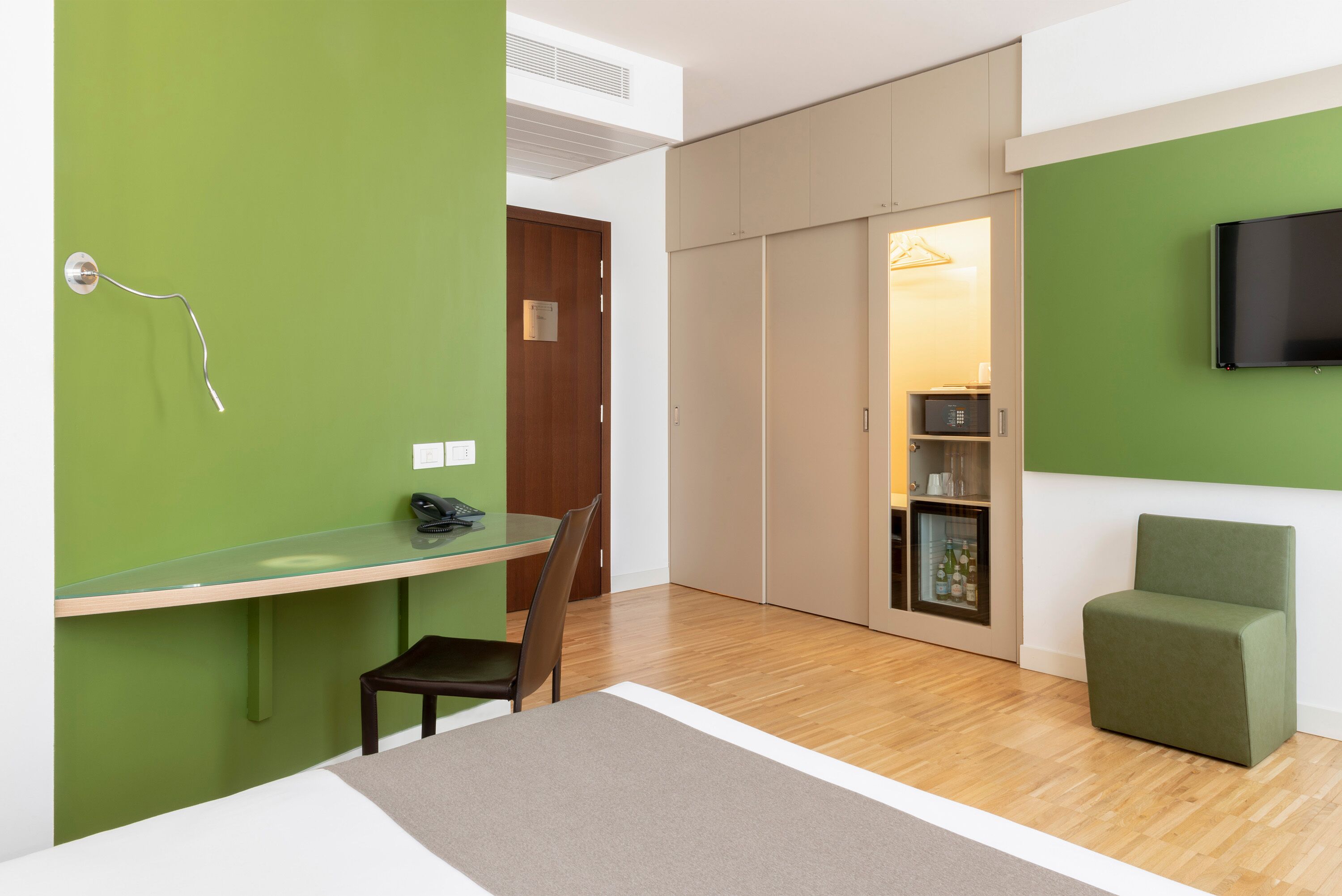 A modern hotel room with a glass desk, a green wall, a wooden door, a wardrobe, a green armchair, and a flat-screen TV.
