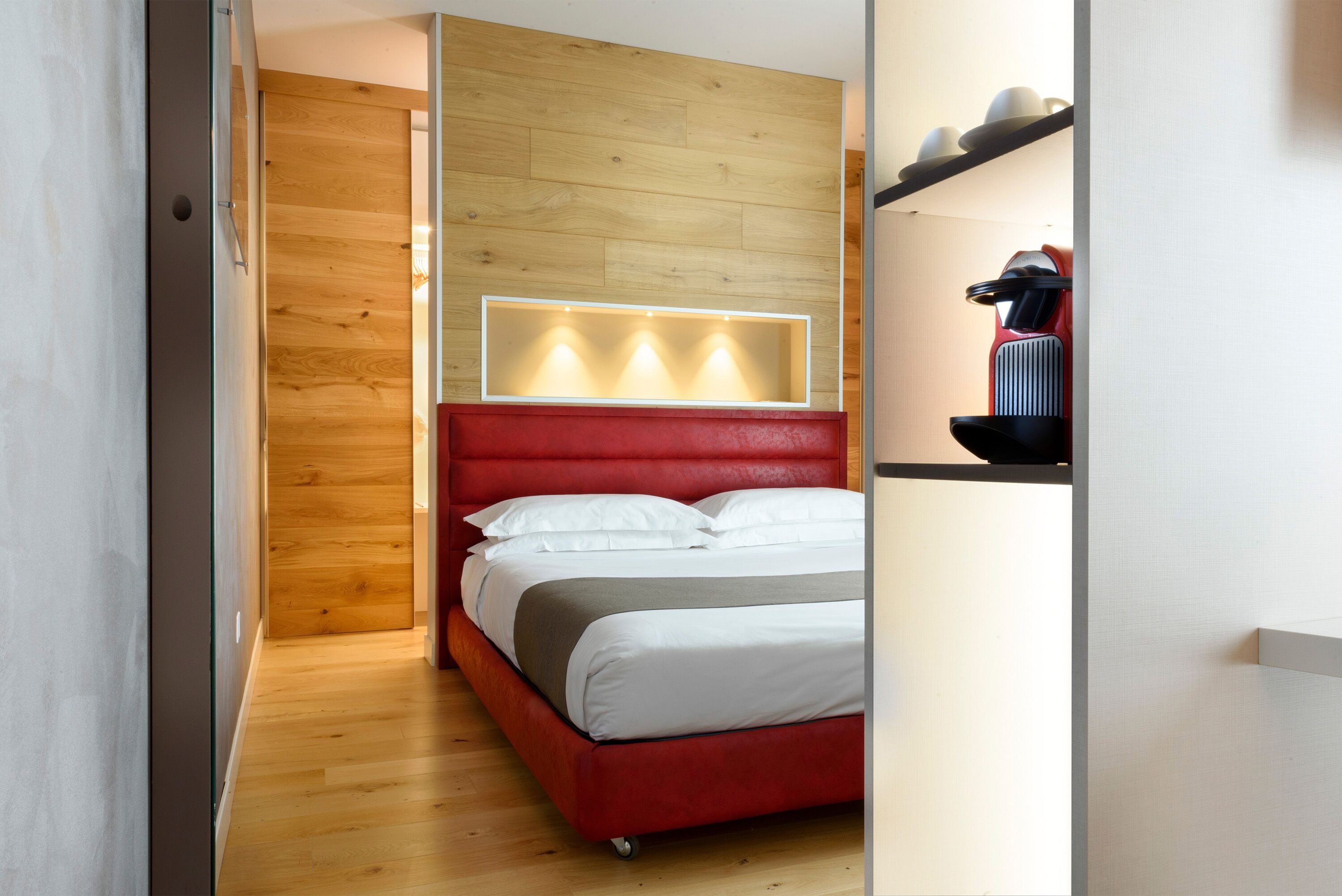 A narrow, modern hotel room with a wooden wall, a red headboard, and a built-in light panel above the bed.