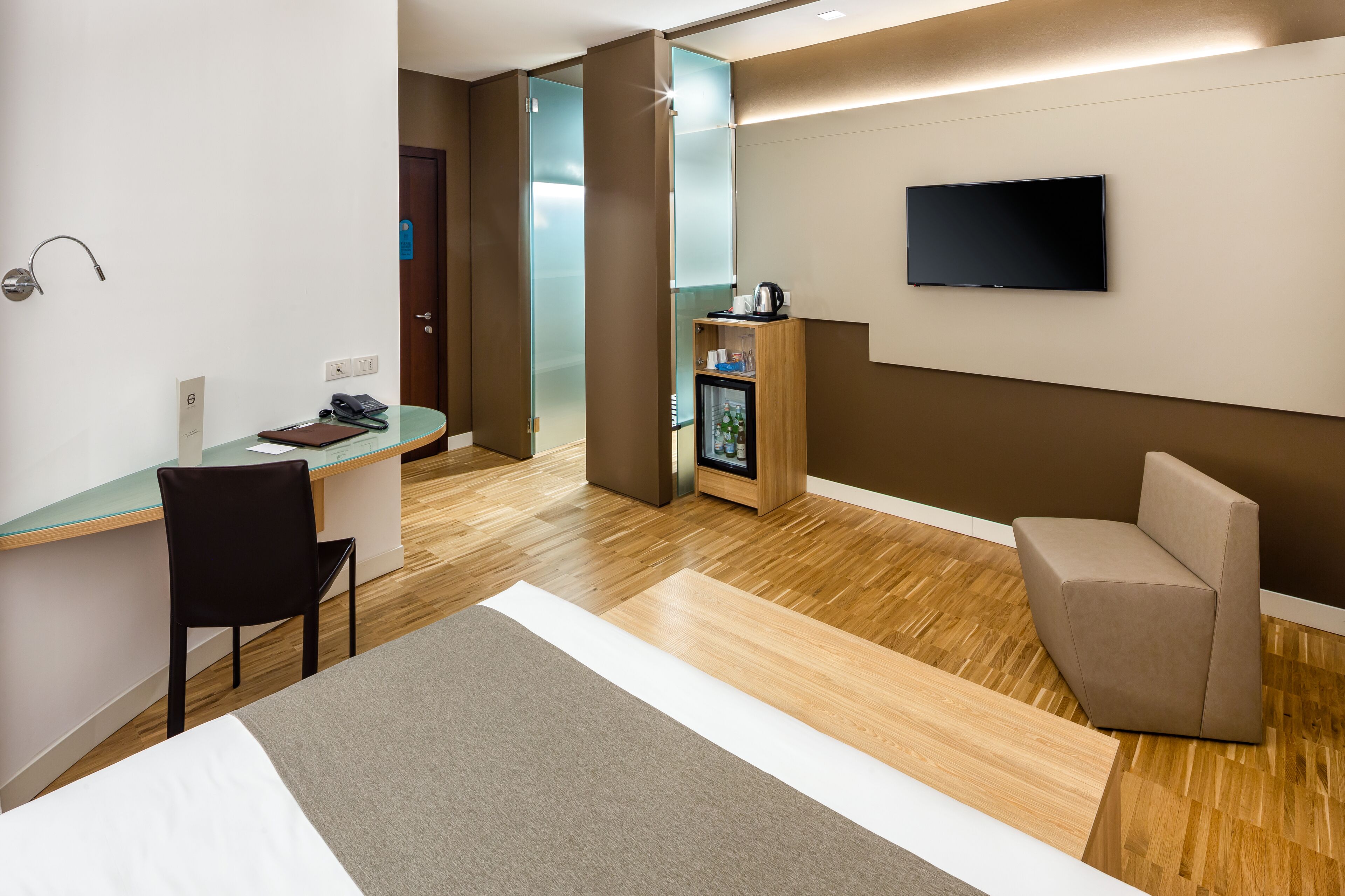 Camera d'hotel moderna con pavimento in legno, un letto, una scrivania con sedia, una televisione e un minibar.