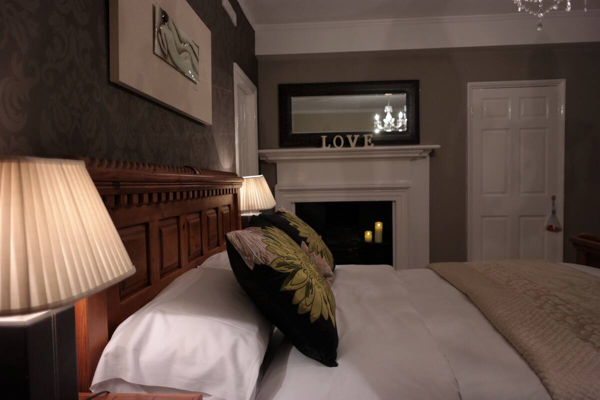 A neatly made bed with a floral pillow, a white lamp, and a fireplace with the word "LOVE" above it.