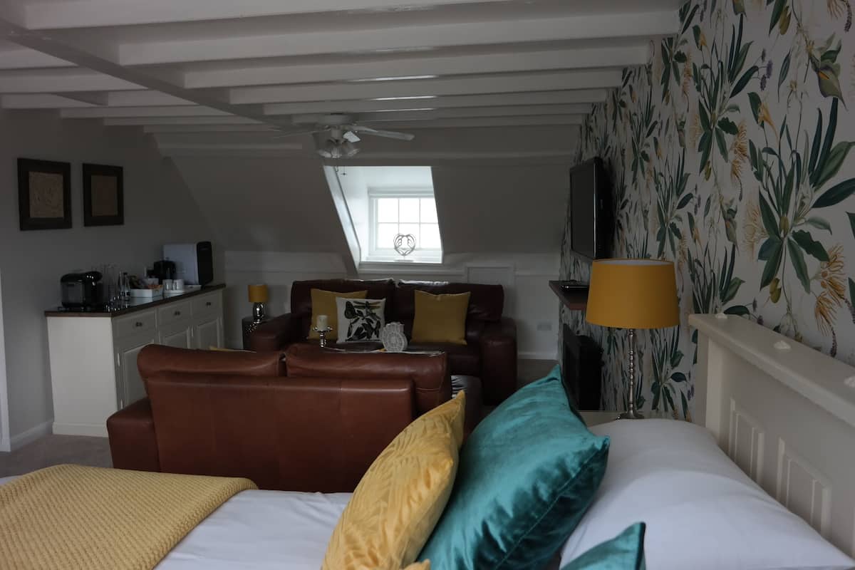 A bedroom with a loft-style ceiling, a bed with multiple pillows, a brown leather sofa, a small desk with a lamp, and a window with a view of the sky.