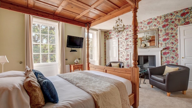 A four-poster bed with a wooden headboard, a television mounted on the wall, a fireplace with a mirror above it, and a floral wallpaper. - White Linen Whitby, Room Only (Whitby)
